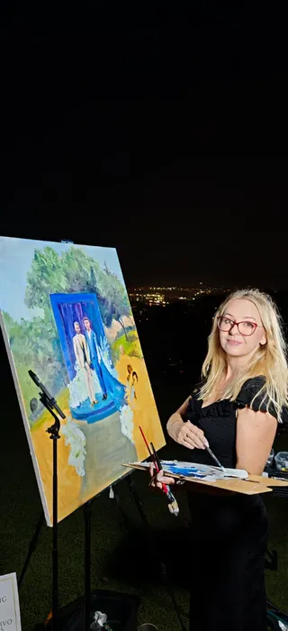 Live wedding painter Marta with her palette beside a finished couple portrait, overlooking the city lights at a hilltop wedding